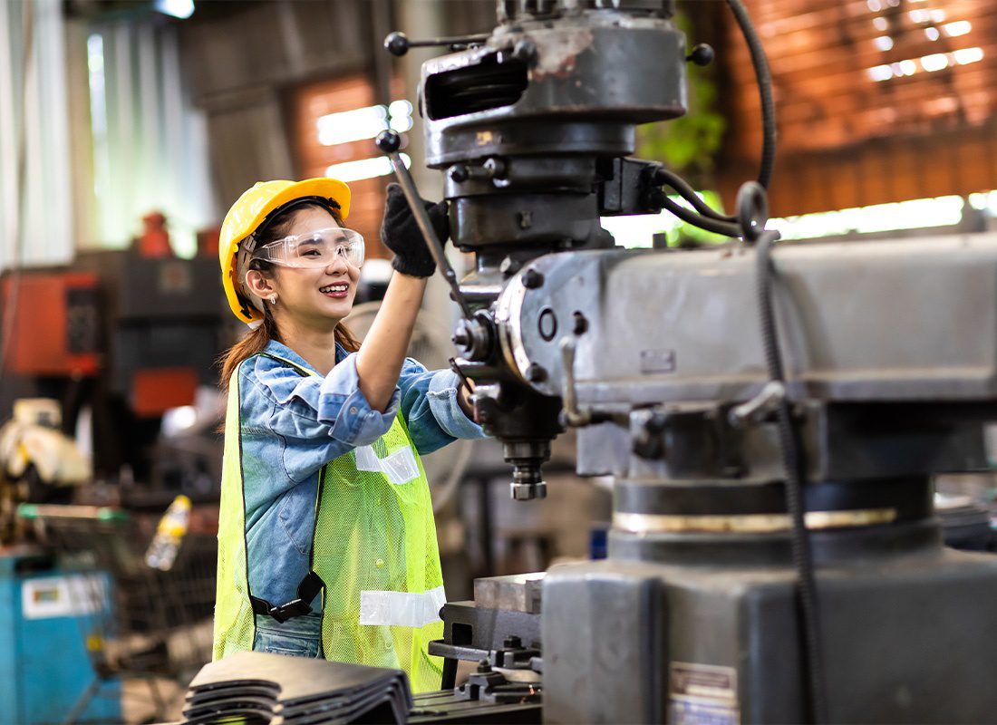 Insurance by Industry - Happy Female Manufacturing Worker Wearing Safety Goggles While Using a Control Lathe Machine to Drill Components in a Warehouse
